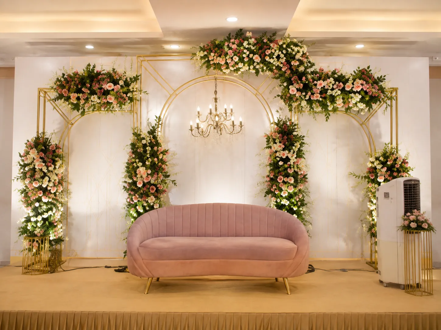 Reception stage with a blush pink settee, white floral arches and chandelier in the air-conditioned hall