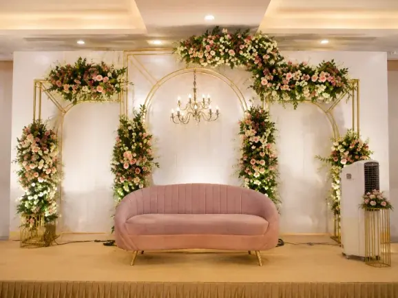 Reception stage with a pink settee framed by white floral arches and a chandelier